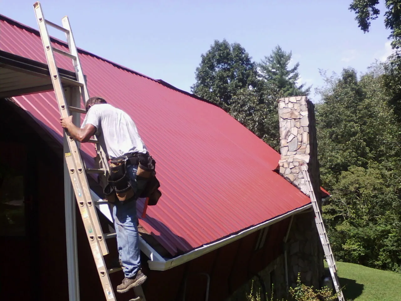 Roofer on a ladder beside a red metal roof and stone chimney — Soffit & Fascia Repair by Roofing Brothers in Harris